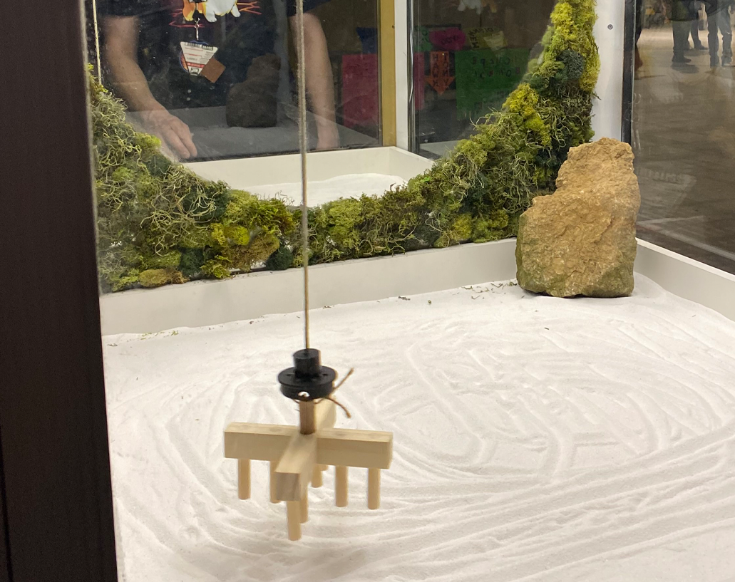 a wooden rake on a claw machine string dangled over the sand covered base of a claw machine